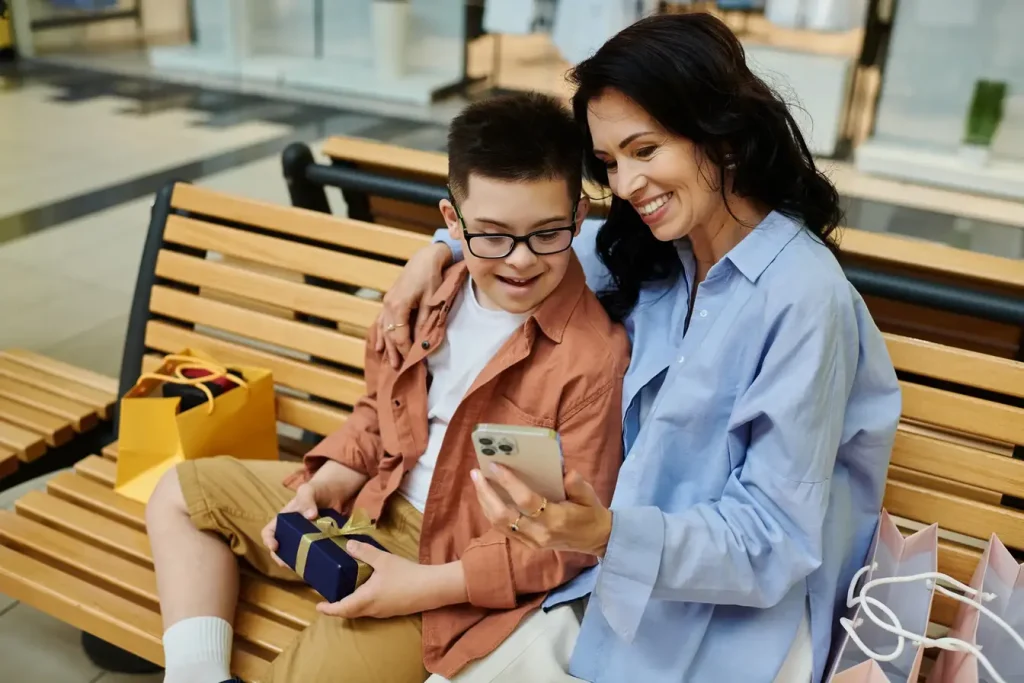 Woman and young boy with disability smiling while sitting on a bench outside and looking at phone